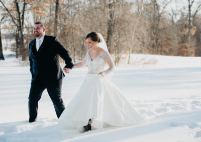 A groom in a black suit and a bride in a white dress and veil walk in the snow