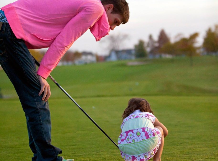 A father and young daughter on a golf course. The father holds a club and bends over to look at his daughter, who crouches down.