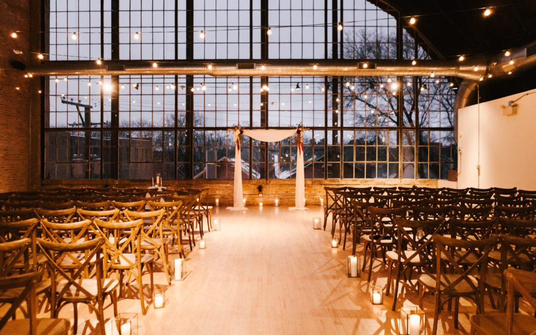 Chairs set up in front of an altar for a wedding. There is a soft glow from candles and lights strung from the ceiling. Behind the altar is a wall made of window. Outside is a winter scene.